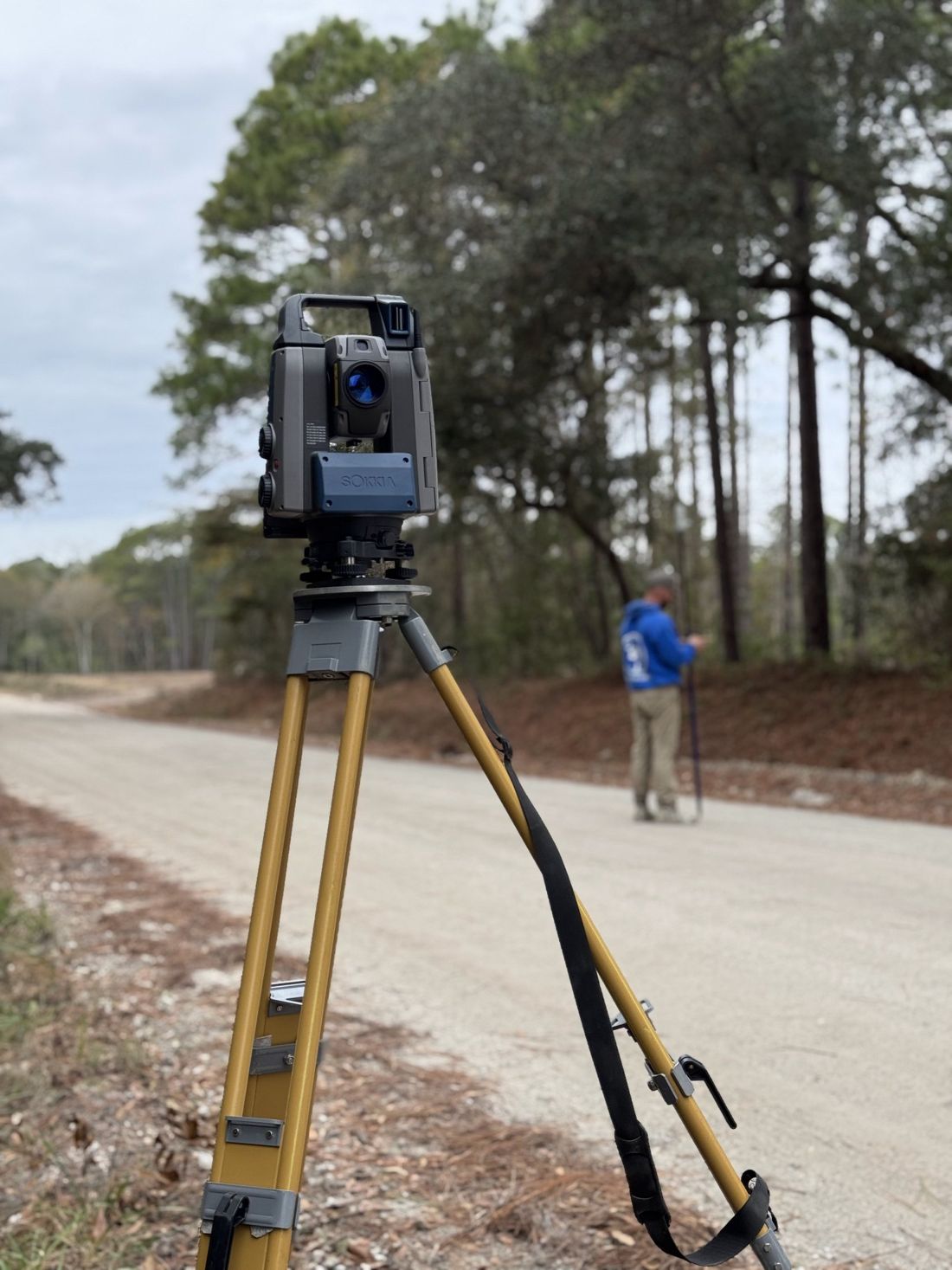 A CMLS crew member holding a prism pole on a dirt road while a Sokkia robotic total station on a yellow tripod tracks him from the foreground